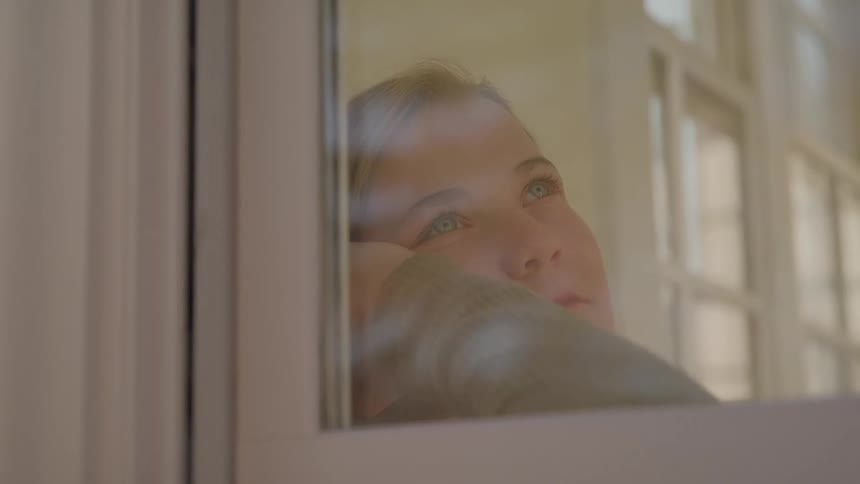 A young woman stands beside the window at midday staring out with a sad expression