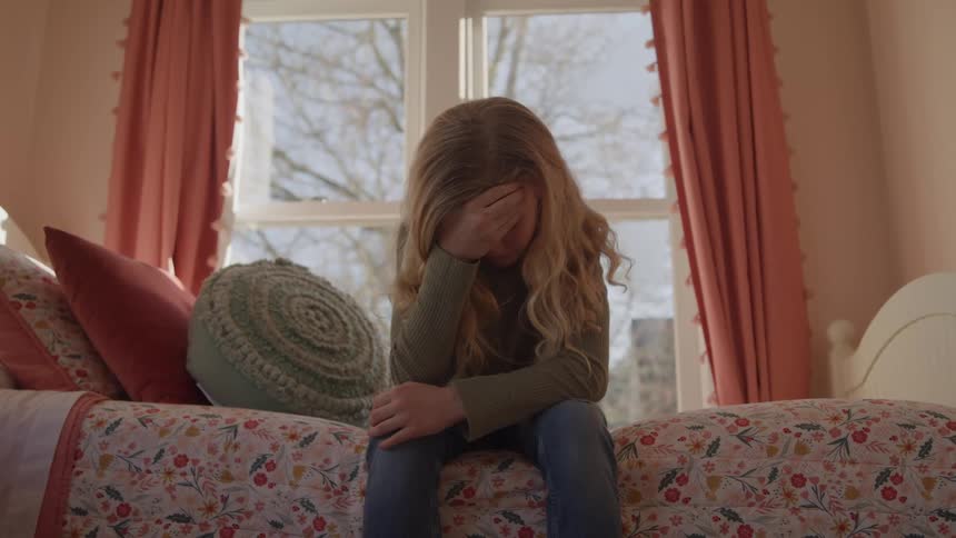 A young girl with long blonde hair sits on her bed and buries her face in her hands looking upset