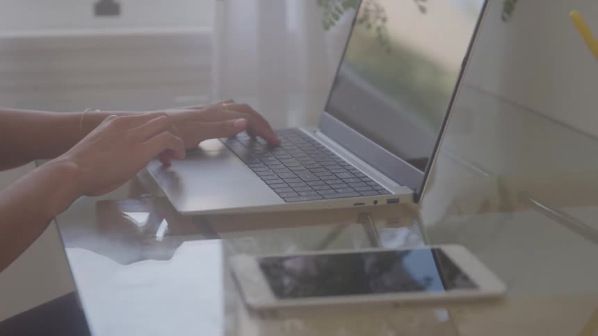 A persons hands rapidly typing on a laptop keyboard with a smartphone lying nearby on a transparent table
