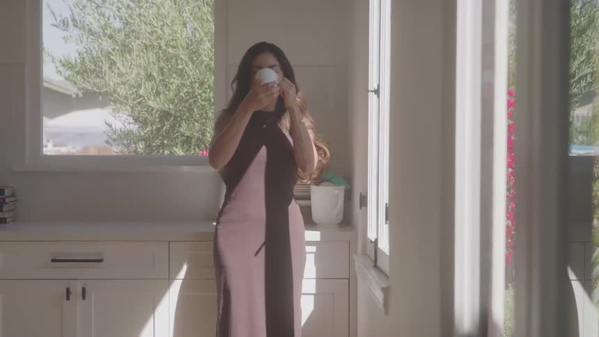 Woman in sleeveless dress holds white mug in sunlit kitchen with garden view through window