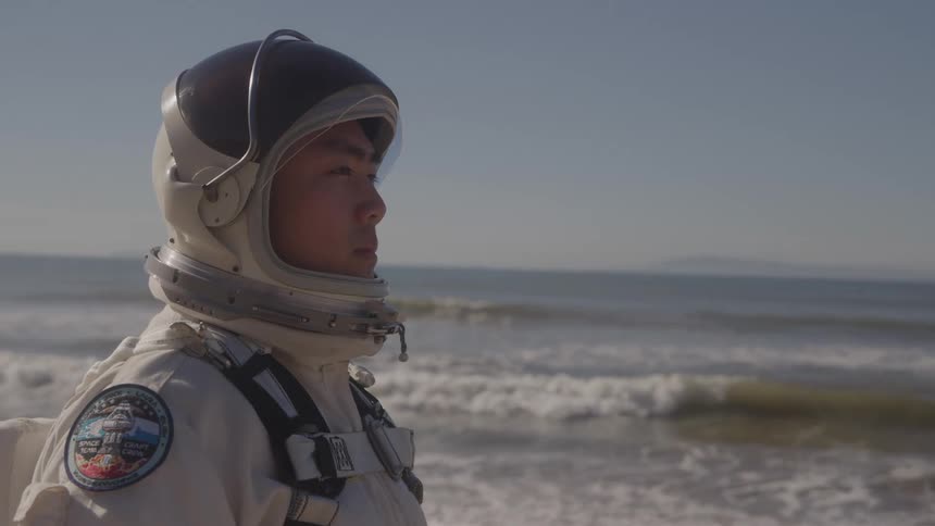 Man in spacesuit gazes at horizon on beach with waves in background
