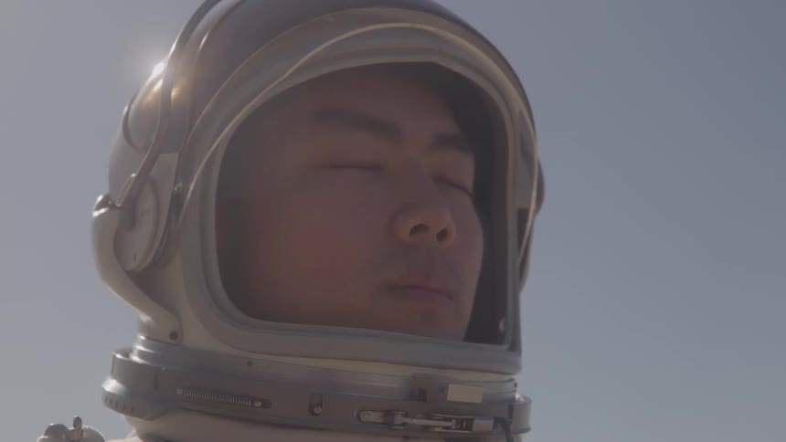Man wearing space helmet stands with eyes closed under blue sky in outdoor setting