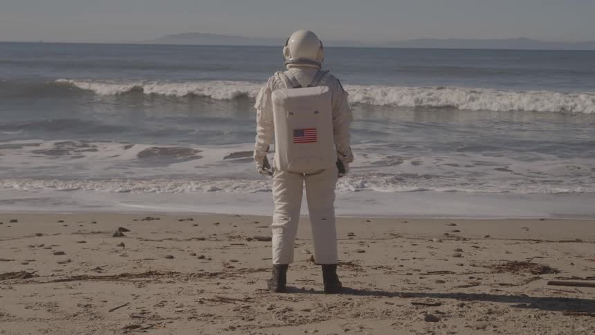 A person wearing a white spacesuit with a helmet and carrying a backpack with an american flag displayed behind them stands on the beach sand on a sunny day watching the ocean waves hit the shoreline