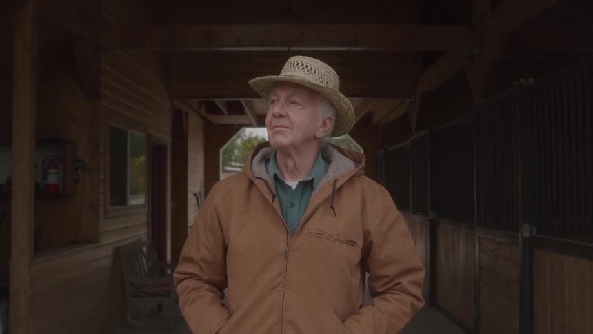 The man is standing outside the stable with his hands in his pockets