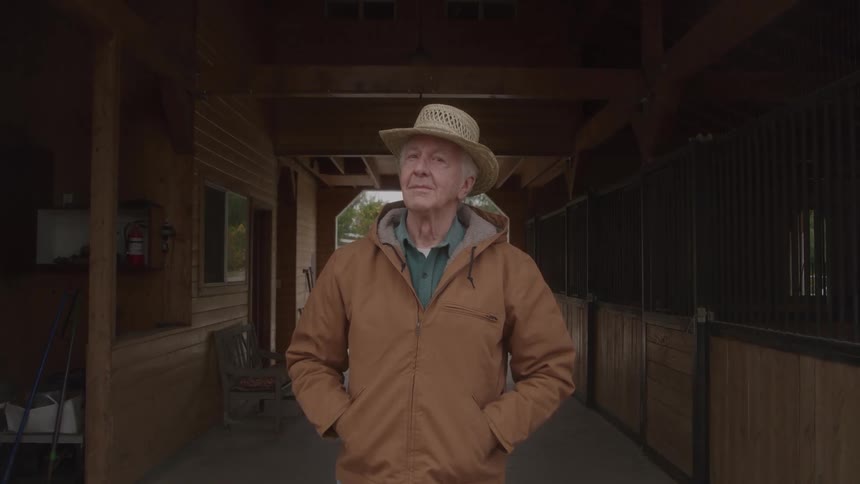 An older man stands in a stable doorway with his hands in his pockets gazing calmly with a quiet steady expression