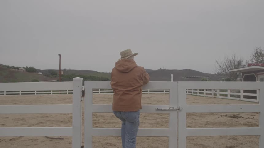 Man wearing a straw hat leaning on a white fence observing an open area