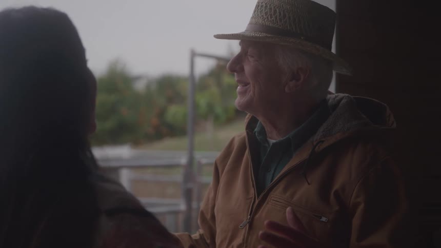 An older man in a straw hat and brown jacket smiles and talks to a person whose back is to the camera standing in a doorway overlooking a rural scene