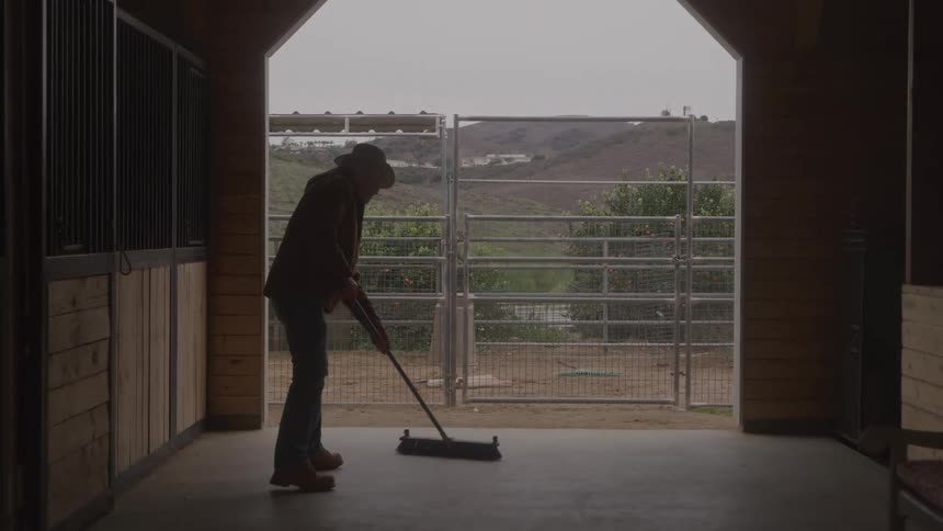 Man sweeping the floor of a stable with hills visible in the background