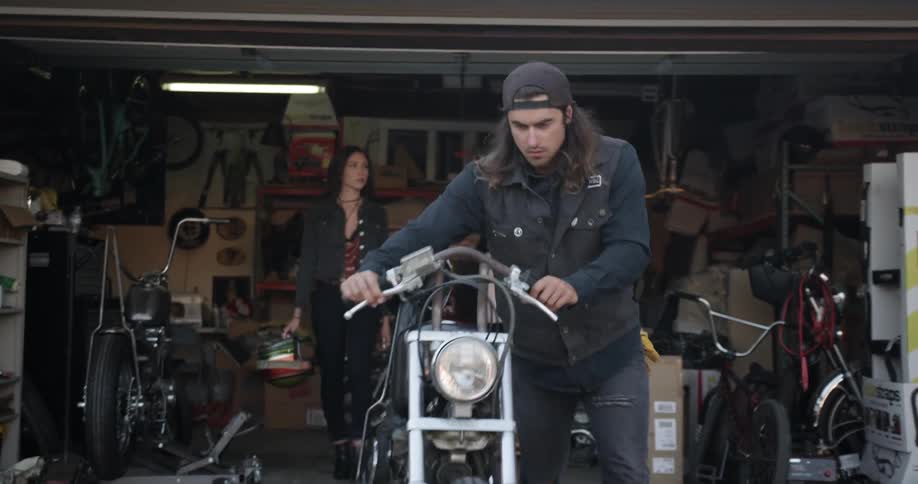 A man rolls his motorcycle out of a garage while a woman walks out behind him