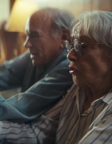 Two older people sit next to one another in an indoor space and react to something