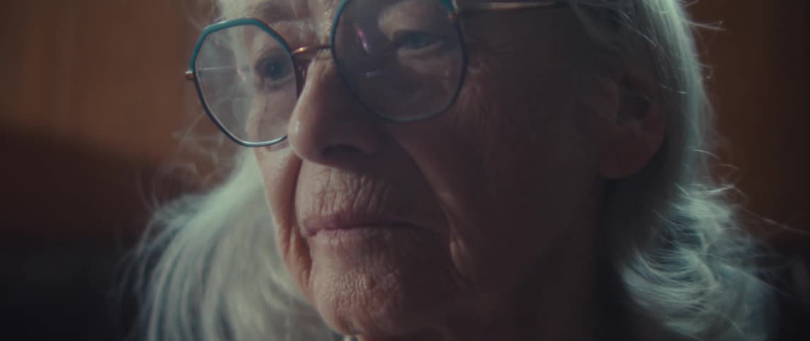 A close view shows the face of an older woman with gray hair and large bluerimmed glasses with a serious expression in low indoor light