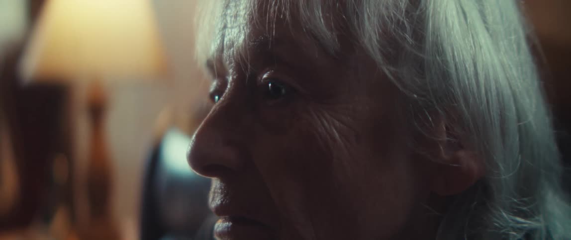 A close side view shows an older woman with bright eyes and gray hair watching something intently in a dimly lit room with a warm lamp in the background
