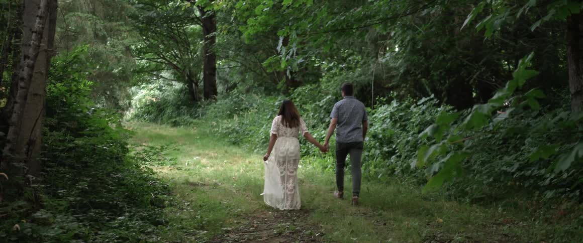 A man and woman hold hands while walking through a forested area