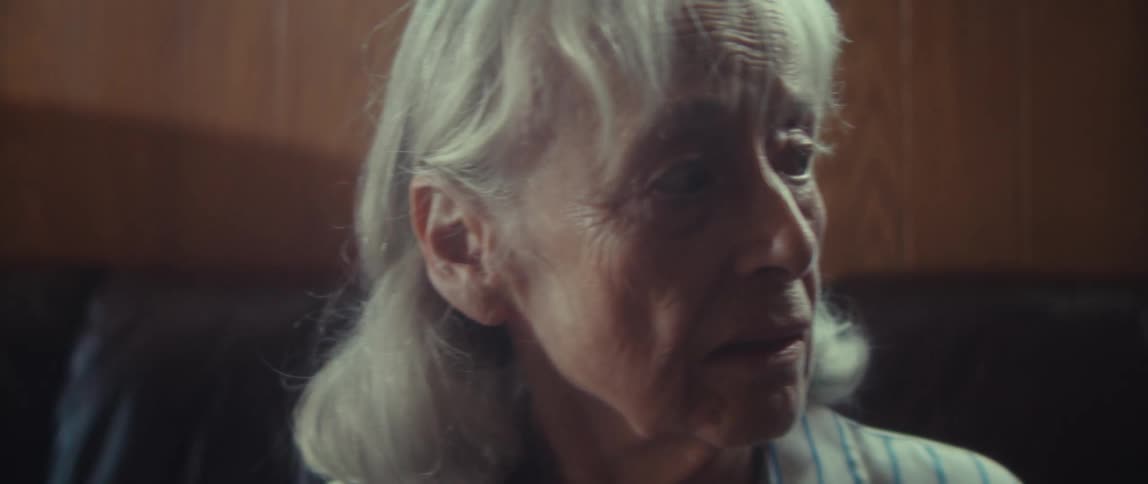 A close view shows the face of an elderly woman with white hair and a striped shirt looking intently toward the right side of the dark warmtoned room