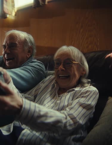 An elderly couple sitting on a couch laughing and moving back and forth as they hold out console controllers while playing together