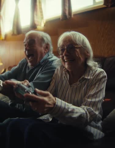 The video is a mediumwide shot of an elderly couple intensely playing a video game on a console sitting together on a couchthey are both laughing and shouting excitedly while holding controllers with the woodpaneled wall and windows visible