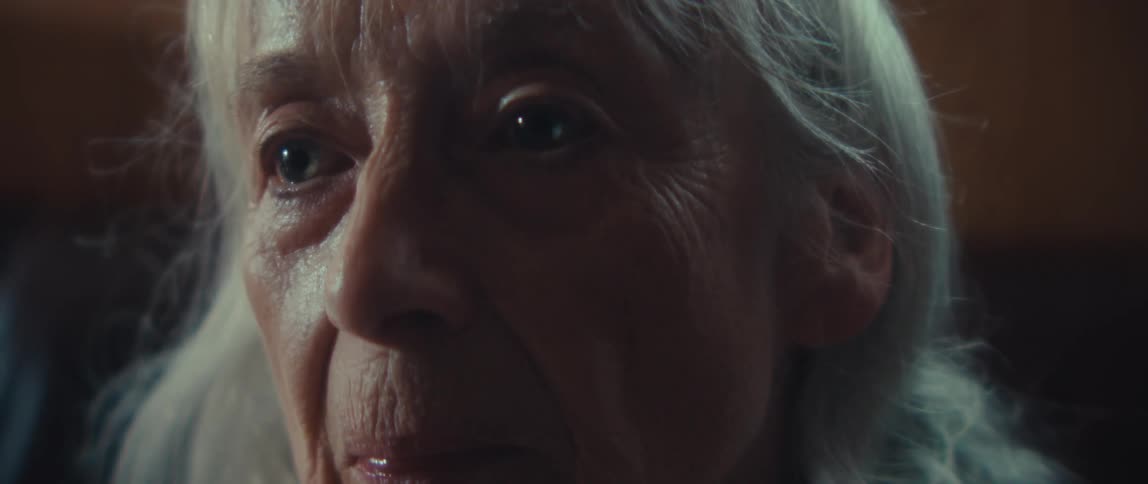 A close dark view shows an elderly woman with white hair removing her large teal and goldrimmed glasses in a warmly lit intimate room