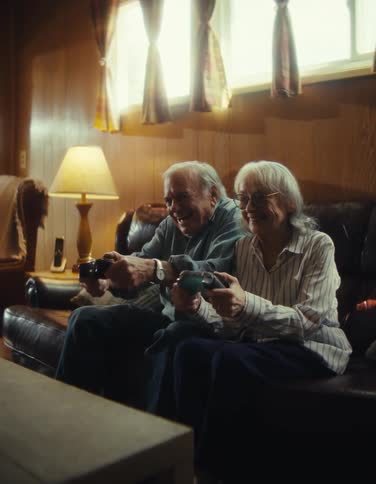 An elderly couple sitting on a couch laughing as they move around console controllers as if playing together
