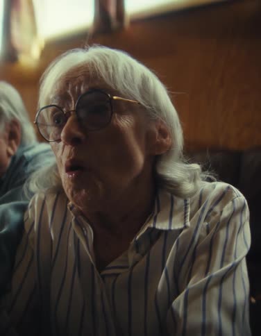 A smiling elderly couple sits close together in a sunny living room the woman holding a video game controller as they both cheer and react excitedly to the screen