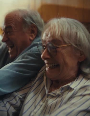 A joyful older woman in a striped shirt and glasses laughs alongside an older man in a blue shirt as they sit close together on a couch playing a video game