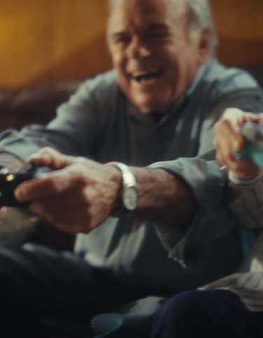 An elderly couple sitting on a couch laughing as they move around console controllers while playing together