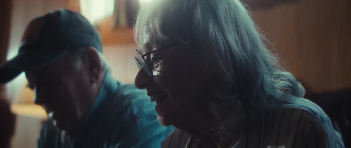 An elderly woman laughs warmly her face lit by soft light as she shares a moment with a man beside her