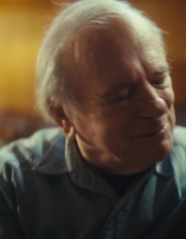 A close up shot focuses on a joyful elderly woman in glasses then pans to her equally happy male companion as they watch something off camera