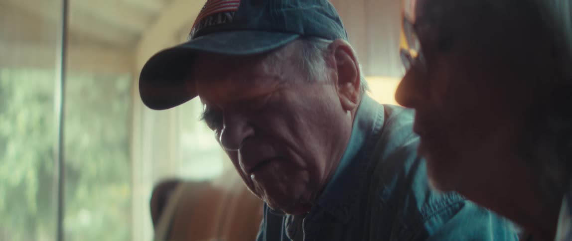 An older man wearing a ball cap and a collared shirt positioned next to the partial profile of a older woman