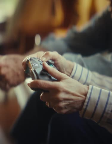 A close view shows two people sitting on a couch holding and playing with video game controllers in a warm dimly lit living room
