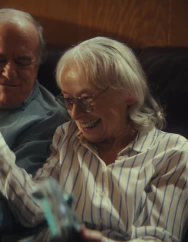 The video is a closeup shot of an older couple laughing joyfully while seated indoorsthe man on the left is laughing heartily and the woman on the right wearing glasses and a striped shirt is holding a remote control and smiling widely