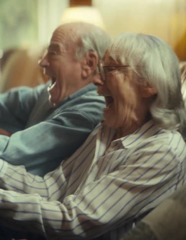 An elderly man and woman screaming and making happy faces while playing video games with controllers in a living room during daytime