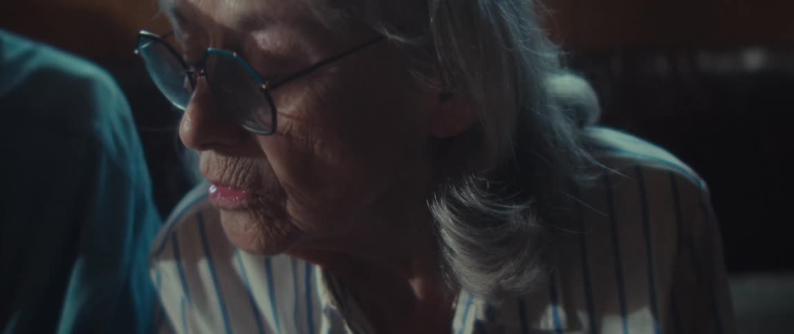 The video is a closeup shot of two elderly people looking down at something offcamera in a dim settingthe man on the left is wearing a veteran baseball cap and a blue collared shirt while the woman on the right is wearing glasses and has w
