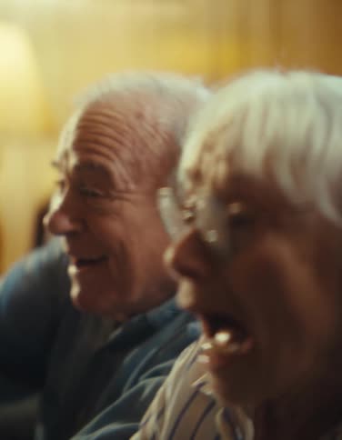 An older woman wearing a striped shirt and glasses laughs happily while sitting next to an older man who is also laughing with his mouth wide open