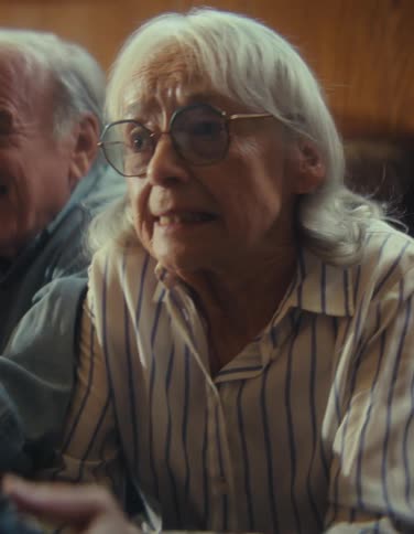 The video is a mediumclose shot of an elderly couple seated closely together passionately engaged in playing video gamesthe man on the left is cackling with laughter while the woman on the right wearing glasses and a striped shirt is foc