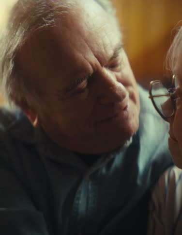 A close view shows a happy older man and woman with gray hair smiling and leaning their heads close together in soft warm indoor light