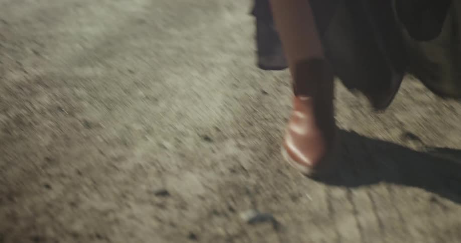 A woman wearing a dress and ankle boots kicks up dirt as she walks