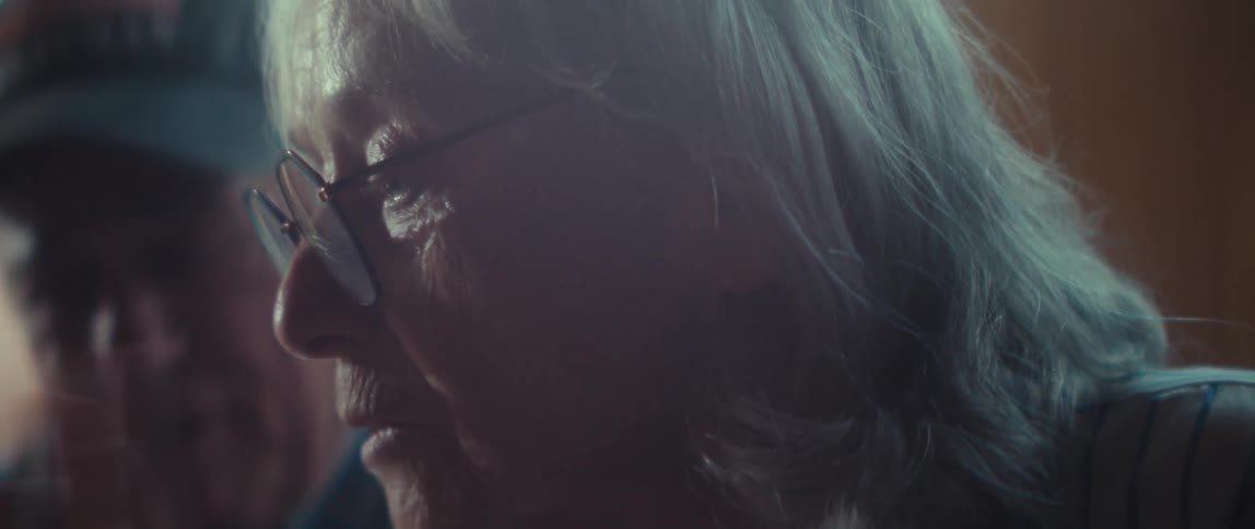 A close warmtoned view shows a senior man in a blue veteran baseball cap leaning down as he speaks closely with an older woman in glasses