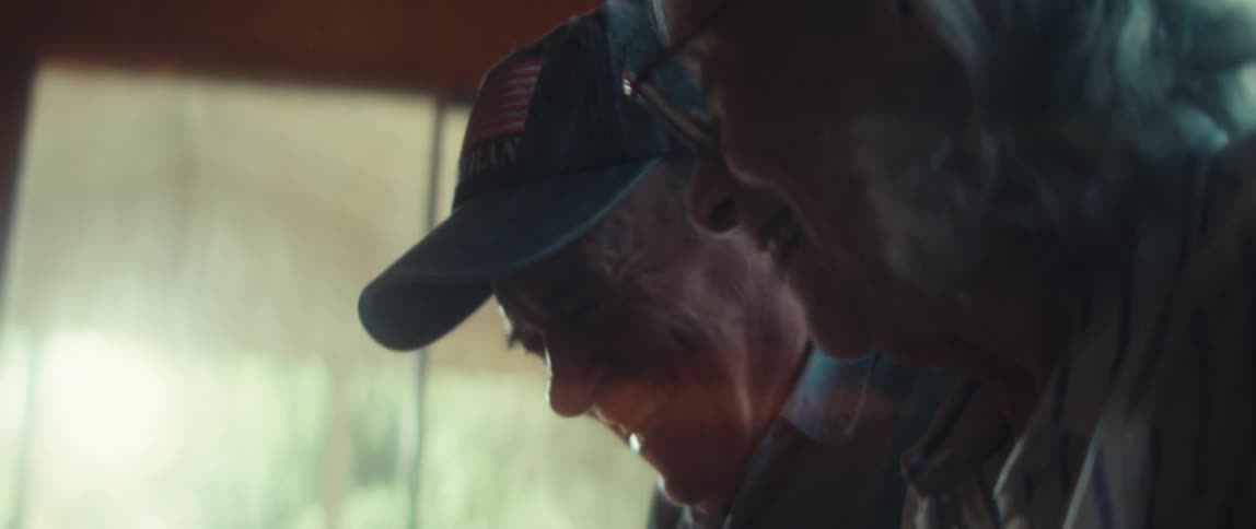A close side view shows an older man wearing a dark cap smiling while an older womans face and glasses are visible nearby in soft indoor lighting