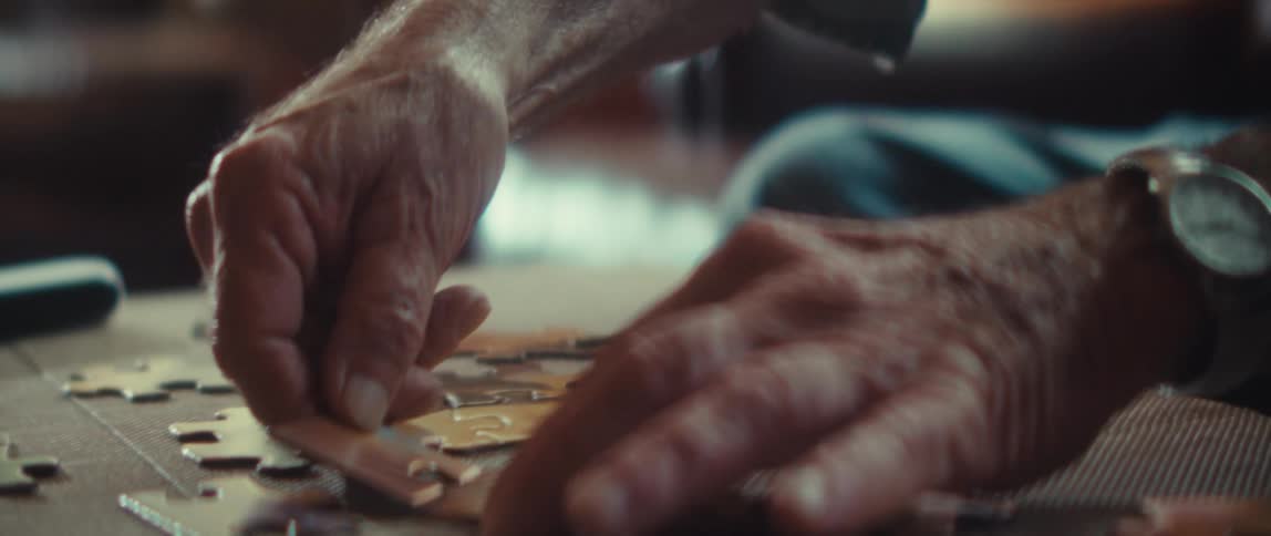 A close view shows the hands of a man working on a jigsaw puzzle picking up a puzzle piece and then the mans face appears as he focuses in a warmly lit indoor space
