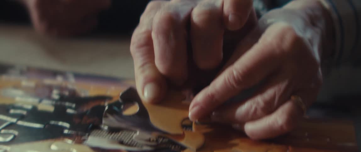 The video is an extreme closeup of a persons hands working on a puzzle on a wooden tablethey are holding and carefully fitting a small white puzzle piece into the rest of the image