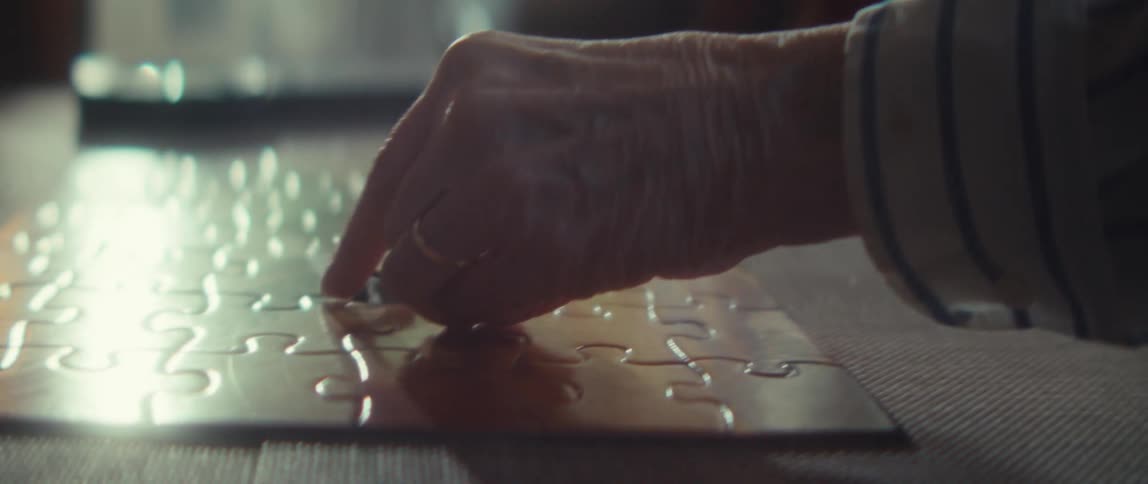 A close view shows the wrinkled hand of an older person completing a shiny silver jigsaw puzzle on a dark table with two blurry people in the background