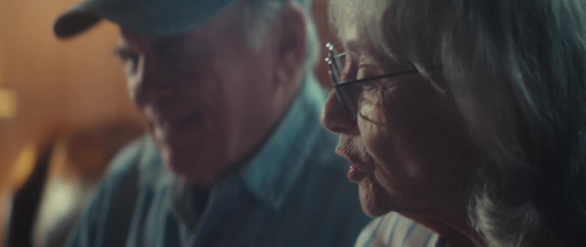 An elderly couple sit closely at a table in a warm dimly lit room working on a jigsaw puzzle