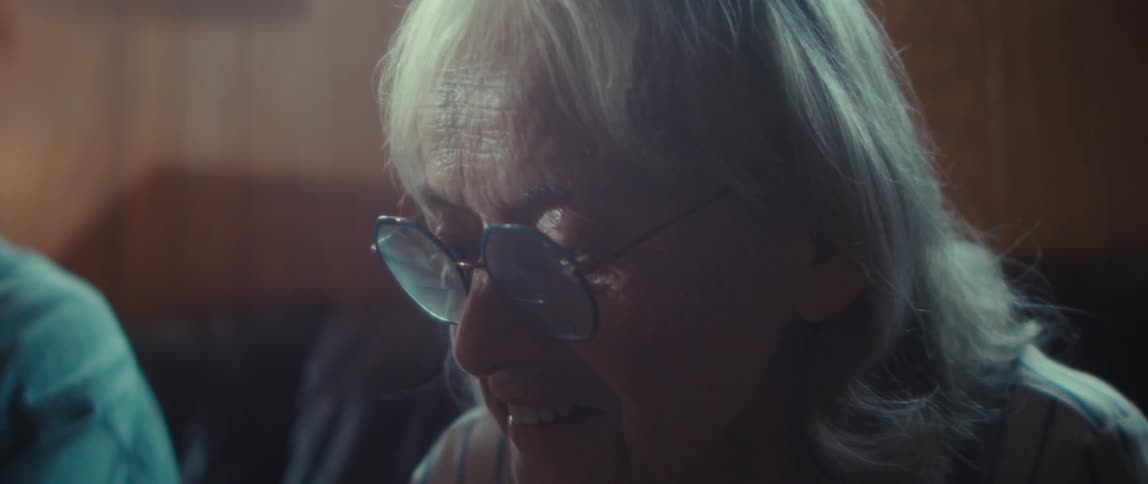 An elderly couple sitting side by side on a couch with him turning his head to ask her a question and she smiles then laughs as she answers