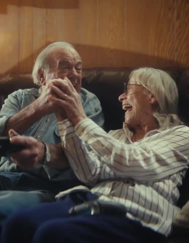 An elderly couple sits on the couch in the living room at midday both overly excited as they play a video game with controllers