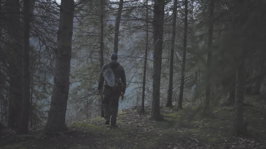 A man is walking through a forest and also he has a fishing rod in his hand and he has a bag and a cap on his head