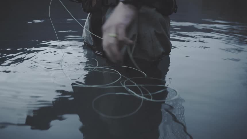 Man standing waistdeep in dark water calmly coiling fly line with focused patient attention