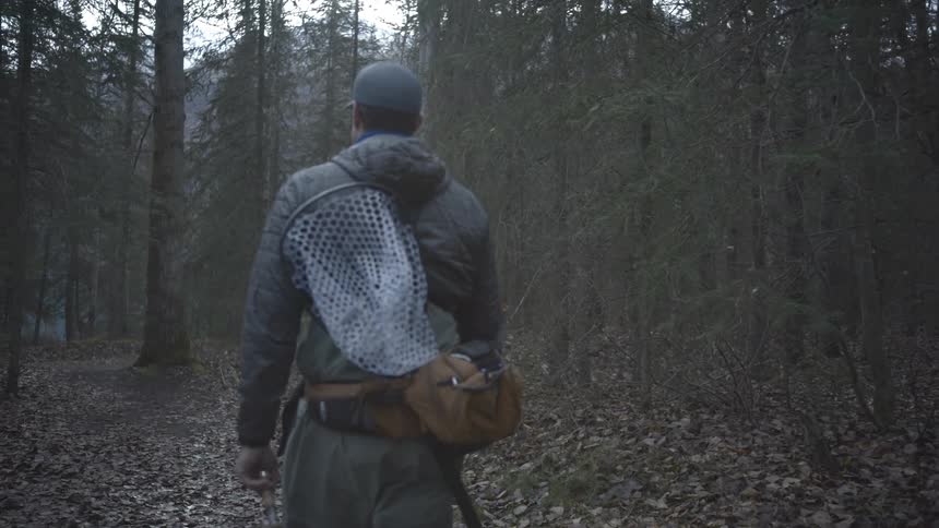 The man is hiking through the forest carrying fishing gear