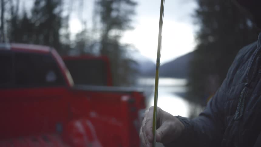 The hands of a man in a quilted jacket assemble a fishing rod near the tailgate of a red pickup truck with a lake and mountains visible in the misty background