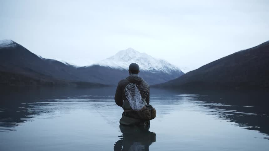 Fisherman kneeling in a calm lake surrounded by snowy mountains