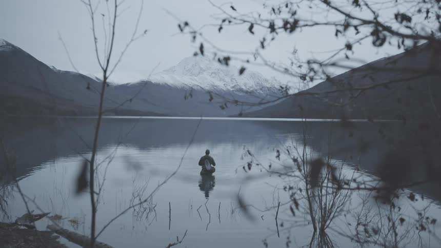 Person fishing in a calm lake surrounded by snowcapped mountains